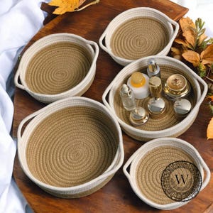May include: Set of five round, woven rope baskets with white trim. The baskets are arranged in a circle on a wooden surface. The largest basket is in the centre and contains several small bottles.