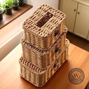 Puede incluir: Tres cajas de almacenamiento de mimbre tejido apiladas sobre una superficie de madera. La caja superior tiene una abertura rectangular. Cada caja tiene un cierre de palanca oscuro. Las cajas son de color marrón claro.