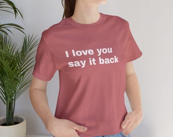 May include: A dusty rose short-sleeved t-shirt featuring the white text "I love you say it back" printed across the chest. The shirt has a crew neckline and a standard fit, paired with blue denim jeans, showcasing a casual and simple design.
