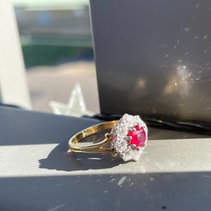 Könnte beinhalten: Ein goldener Ring mit einem großen roten Rubin, der von kleineren Diamanten umgeben ist.