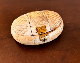 May include: An oval-shaped, off-white box with a hinged lid and a decorative brass clasp. The box is made of small, rectangular pieces, with carved geometric and sunburst patterns. The box is resting on a dark brown wooden surface.