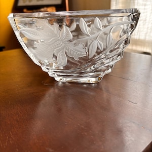 May include: A clear glass bowl with a floral leaf design etched into the side. The bowl has a rectangular shape with angled sides and sits on a dark wooden surface. The background includes a framed sign that says "Blessed".