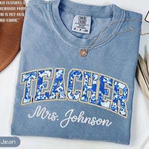 Puede incluir: Camiseta azul vaquero con la palabra "TEACHER" en un diseño floral, delineada con pequeñas cuentas doradas. Debajo, "Mrs. Johnson" en cursiva. La camiseta tiene una etiqueta Comfort Colors.