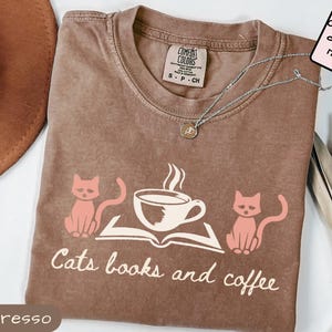 Puede incluir: Camiseta color espresso con un gráfico de dos gatos rosas, una taza de café y un libro abierto. El texto "Cats books and coffee" está impreso debajo. También se ven un collar plateado y una etiqueta Comfort Colors.