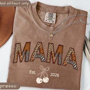 Puede incluir: Camiseta color espresso con la palabra "MAMA" en un diseño audaz de estampado animal. La camiseta incluye el texto "Est. 2026" y un gráfico de cerezas. Un collar plateado con un colgante está sobre la camiseta.