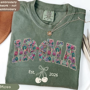 Puede incluir: Camiseta verde musgo con la palabra "MAMA" en un diseño floral, un lazo y el texto "Est. 2026". Un collar dorado con un colgante descansa sobre la camiseta. La camiseta está etiquetada como "Comfort Colors" y "S-P-CH".