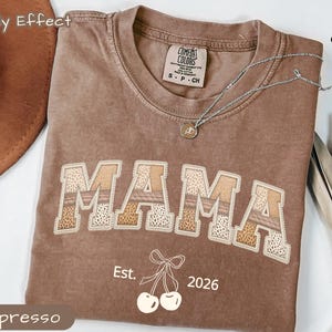 Puede incluir: Camiseta color espresso con la palabra "MAMA" en un diseño de patchwork. La camiseta presenta el texto "Est. 2026" y un gráfico de cerezas. Un collar plateado con un colgante está drapeado sobre la camiseta. La camiseta está etiquetada como "Comfort Colors".