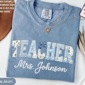 Puede incluir: Camiseta azul vaquero con la palabra "TEACHER" en un diseño de patchwork y "Mrs. Johnson" bordado debajo. Un collar dorado con un colgante y una etiqueta que dice "Comfort color 1717" están en la camiseta.
