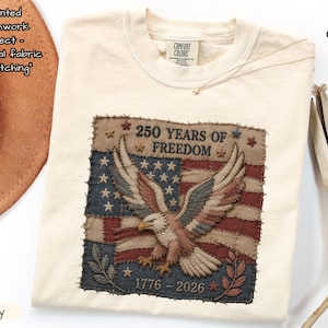 May include: Ivory-coloured t-shirt with a printed patchwork design of an American flag and an eagle. The text reads "250 YEARS OF FREEDOM 1776-2026". The shirt is a Comfort Colors brand.