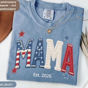 Puede incluir: Camiseta azul vaquero con la palabra "MAMA" en falso bordado. La "M" y la "A" están diseñadas con un patrón de bandera americana roja, blanca y azul. La "M" y la "A" están decoradas con perlas blancas. La camiseta tiene el texto "Est. 2026".