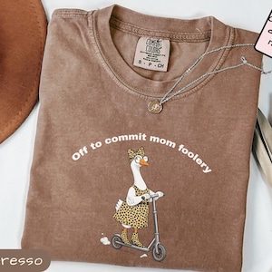 Puede incluir: Camiseta Comfort Colors color Espresso con la frase "Off to commit mom foolery" y un dibujo de un ganso en patinete. El ganso lleva un atuendo con estampado de leopardo. Un collar plateado está sobre la camiseta.