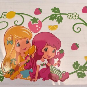 May include: A colorful illustration featuring two cartoon characters with long hair, one yellow and one pink, holding fruit-shaped items. The design includes strawberries, lemons, and a green vine border, creating a cheerful, playful aesthetic.