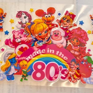 May include: A clear sticker sheet featuring various 1980s cartoon characters including Rainbow Brite, a Care Bear, a Smurf, and Strawberry Shortcake. The central design includes a rainbow with the text made in the 80's and many small colorful stars.