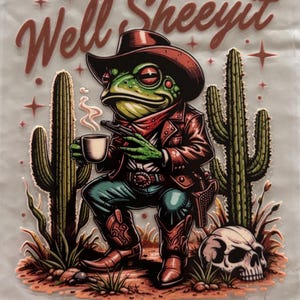 May include: A graphic transfer featuring a green frog dressed as a cowboy in a hat, jacket, and boots, holding a steaming cup and a pistol. The scene includes two cacti, a skull, and the text "Well, Sheeyit" in a brown script font with stars.
