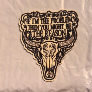 May include: An embroidered patch with a cream-colored background and black stitching. The design features a steer skull at the bottom and decorative scrollwork. The text reads: IF I'M THE PROBLEM THEN YOU MIGHT BE THE REASON, framed by stars.