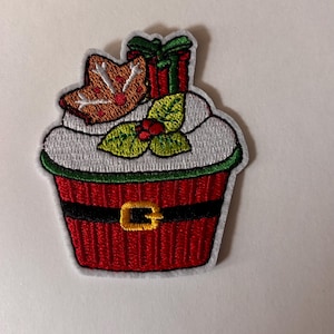 May include: An embroidered patch shaped like a cupcake with a red wrapper, a black belt with a yellow buckle, white frosting, a brown gingerbread cookie, a red gift box with a green bow, and green holly leaves with red berries on top.