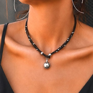 May include: A multi-strand black cord necklace featuring small silver-toned metallic beads spaced along the cords. The necklace includes a central silver-toned spherical pendant suspended from a silver-toned metal loop at the base of the cords.