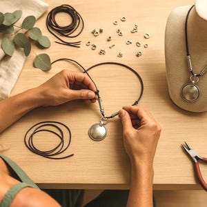 A woman is carefully crafting a necklace using various materials, including leather cords, beads, and pliers, on a wooden table.
