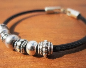 May include: A black leather bracelet with silver beads. The bracelet features a magnetic clasp and several decorative silver beads with various patterns. The beads are round and cylindrical, adding texture to the design.
