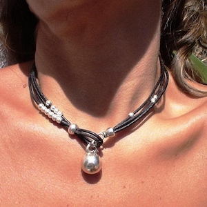 Puede incluir: Un collar de cuero negro con múltiples hebras, adornado con cuentas plateadas y un colgante en forma de campana. El collar se lleva alrededor del cuello. El diseño es sencillo y elegante.