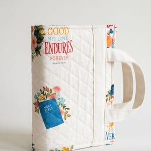 May include: Quilted Bible cover with floral patterns and the text "GOOD HIS LOVE ENDURES FOREVER". The cover has a white background with colorful flowers and a blue patch that says "HOLY BIBLE". It also has a white handle.