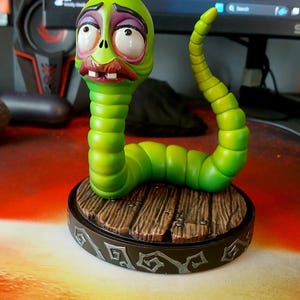 Könnte beinhalten: Eine grüne, cartoonartige Wurmfigur mit einem betrübten Gesichtsausdruck. Der Wurm hat einen segmentierten Körper und ist auf einem runden Sockel mit Holzplankenmuster montiert. Der Sockel hat einen dekorativen Rand.