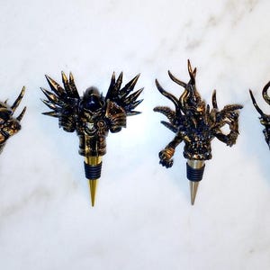 May include: Set of seven gold and black bottle stoppers featuring various fantasy creature designs. Each stopper has a unique design, including a knight, a dragon, a demon, and a griffin.