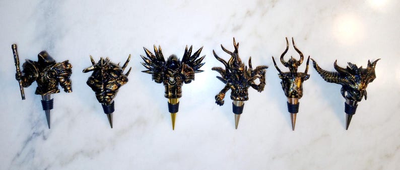 May include: Set of seven gold and black bottle stoppers featuring various fantasy creature designs. Each stopper has a unique design, including a knight, a dragon, a demon, and a griffin.