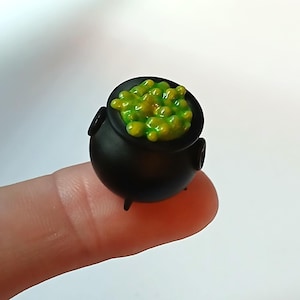 May include: A miniature black cauldron with three small legs and two side handles, filled with a textured, glossy, bright green bubbling liquid substance that appears to be overflowing slightly from the top rim of the round vessel.