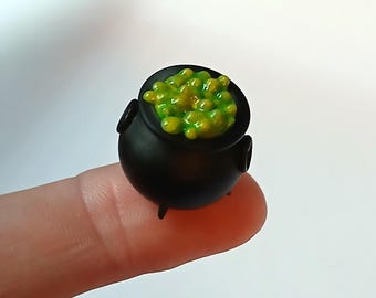 May include: A miniature black cauldron with three small legs and two side handles, filled with a textured, glossy, bright green bubbling liquid substance that appears to be overflowing slightly from the top rim of the round vessel.