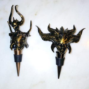 May include: Two gold-coloured bottle stoppers shaped like demonic figures. The stoppers are made of a textured material and have a black rubber base.