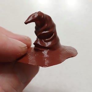 May include: A miniature, dark brown Sorting Hat with a wide brim and a pointed top. The hat is held by a person's fingers. The hat's surface has a textured, ridged appearance.