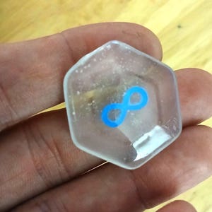 Könnte beinhalten: Eine durchsichtige sechseckige Glasfliese mit einem blauen Unendlichkeitszeichen darauf.