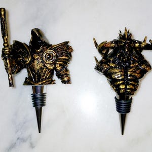 May include: Two gold-coloured bottle stoppers, one featuring a hooded figure with a staff and the other a horned demon. Both stoppers have a black rubber base.