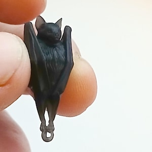 May include: A small, translucent black bat figurine with detailed fur texture on its chest and wings folded against its body. The bat is depicted in a hanging position with a small circular loop at the bottom, designed for use as a pendant or charm.