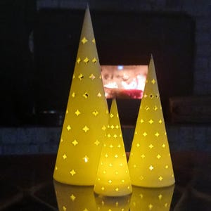 Può includere: Tre luci decorative a forma di cono giallo con ritagli a forma di diamante. Le luci sono disposte in gruppo, di dimensioni variabili, con un bagliore caldo che emana dall'interno. Lo sfondo presenta un'ambientazione scura con un camino.