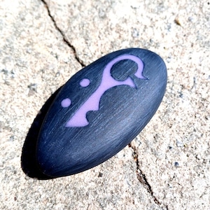 May include: A dark grey stone with a purple symbol carved into it. The symbol is a stylised key with a loop at the top and three dots below it.