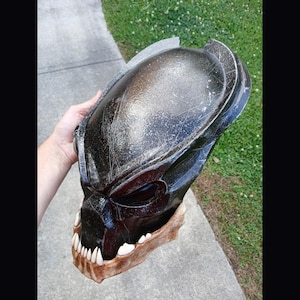 May include: A black and brown Predator mask with white teeth. The mask has a textured surface with silver scratches. The lower jaw is a light brown color. The mask is held by a hand.