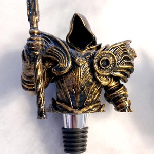 May include: A gold and black metal bottle stopper shaped like a hooded figure holding a staff. The figure is wearing armour and has a dark, hooded cloak.