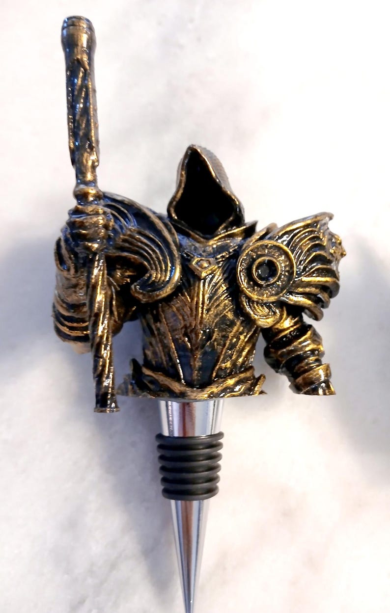 May include: A gold and black metal bottle stopper shaped like a hooded figure holding a staff. The figure is wearing armour and has a dark, hooded cloak.