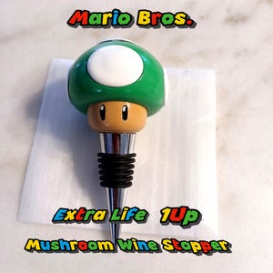 Könnte beinhalten: Ein grüner und weißer pilzförmiger Weinstopfen mit silberner Basis und schwarzem Gummiring. Der Stopfen hat ein Comic-Gesicht mit zwei schwarzen Augen und einem braunen Stiel. Der Text "Extra Life 1Up Mushroom Wine Stopper" ist unter dem Stopfen aufgedruckt.