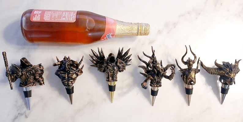 May include: Set of seven gold and black bottle stoppers featuring various fantasy creature designs. The stoppers are made of metal and have a pointed tip for inserting into a bottle.