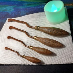 May include: Four miniature wooden brooms with a curved handle and a textured, oval-shaped head. Each broom has a gold-coloured band near the handle. A small, green LED candle sits nearby on a white, textured surface.