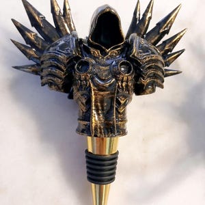 May include: A gold and black metal bottle stopper shaped like a hooded figure with wings. The figure is wearing armour and has a pointed hood. The bottle stopper has a gold metal base and a black rubber stopper.