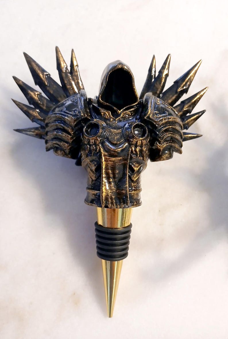 May include: A gold and black metal bottle stopper shaped like a hooded figure with wings. The figure is wearing armour and has a pointed hood. The bottle stopper has a gold metal base and a black rubber stopper.