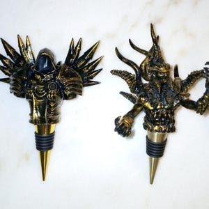 May include: Two gold-painted bottle stoppers, one with a winged, hooded figure and the other with a multi-limbed, horned creature.