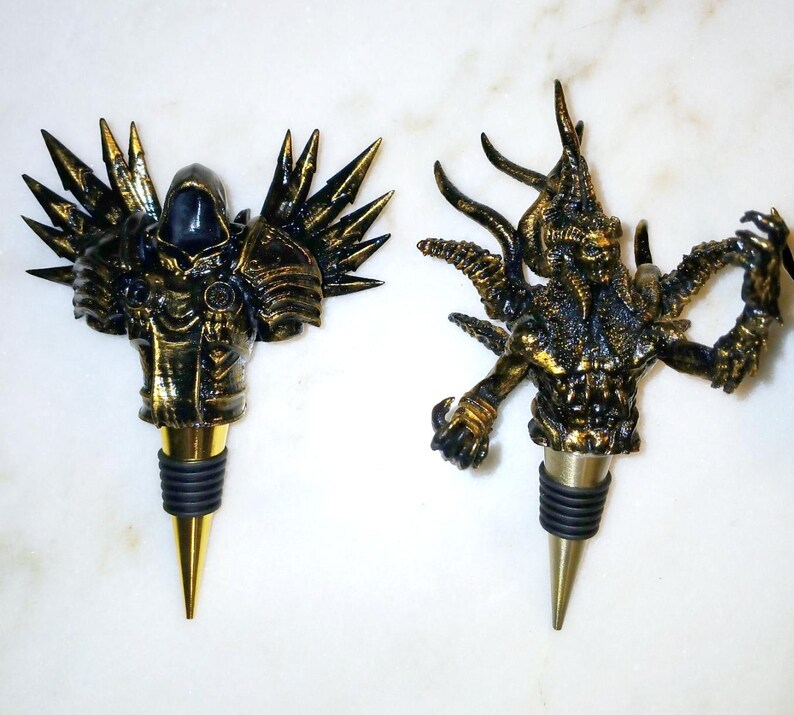 May include: Two gold-painted bottle stoppers, one with a winged, hooded figure and the other with a multi-limbed, horned creature.