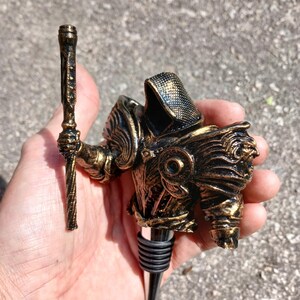 May include: Gold-coloured metal bottle stopper shaped like a knight holding a staff. The knight is wearing armour with a black and gold design.