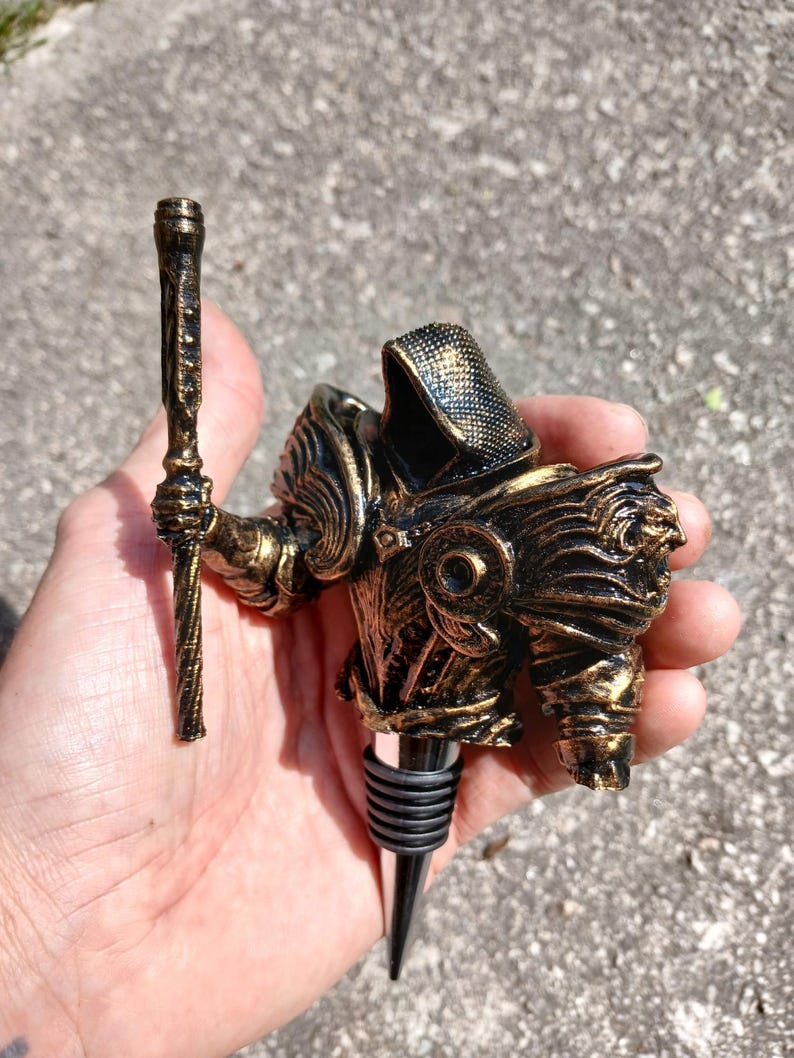 May include: Gold-coloured metal bottle stopper shaped like a knight holding a staff. The knight is wearing armour with a black and gold design.