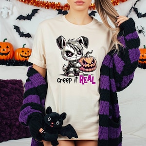 May include: Cream-colored t-shirt with a cartoon bunny holding a jack-o'-lantern. The bunny has a skull-like face and is wearing a black cape. The text "Creep it REAL" is printed below the image. Halloween theme.